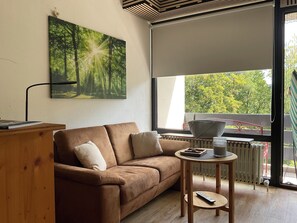 Una sala de estar moderna con un sofá marrón, una pequeña mesita de madera y un ventanal con vistas a árboles.