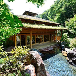 Um edifício de madeira tradicional com telhado de palha, rodeado por um jardim natural com um lago tranquilo.