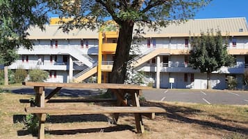 Un edificio de varios pisos con estacionamiento, una mesa de picnic y un árbol al frente.
