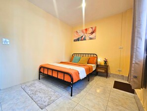 Un dormitorio con una cama, una mesita de noche, una lámpara de pared, un enchufe y una ventana con cortinas.