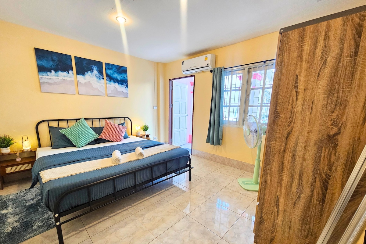 A hotel room with a bed, a nightstand, a wall-mounted air conditioner, a window with curtains, and a view of the outside.