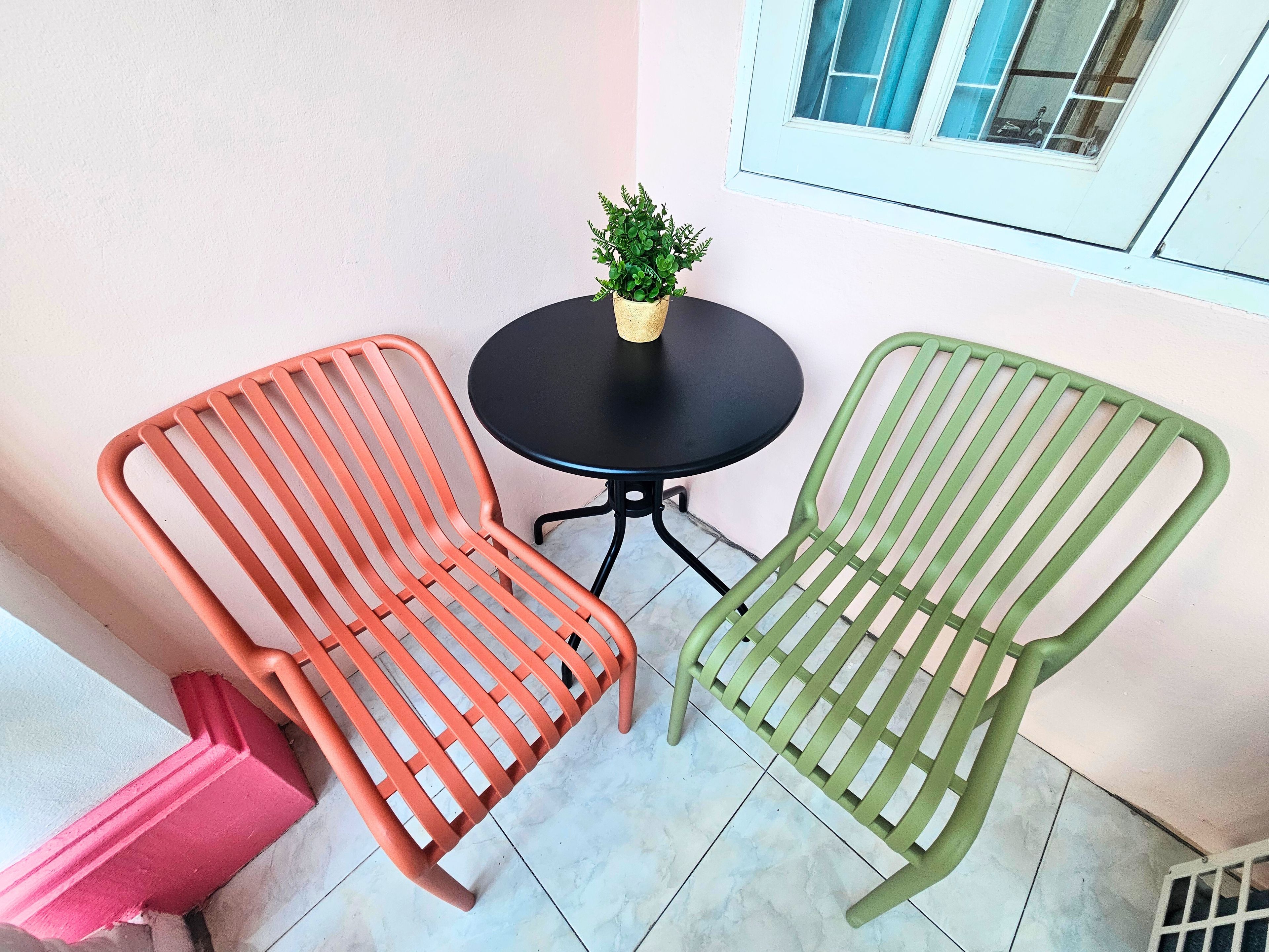Twee metalen stoelen met lattenleuningen, een klein rond tafeltje en een bloempot op een balkon met een witte muur en een raam.