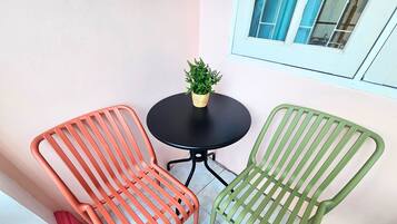 Twee metalen stoelen met lattenleuningen, een klein rond tafeltje en een bloempot op een balkon met een witte muur en een raam.