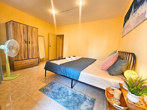 A hotel room with a bed, a fan, a wardrobe, and a painting on the wall.