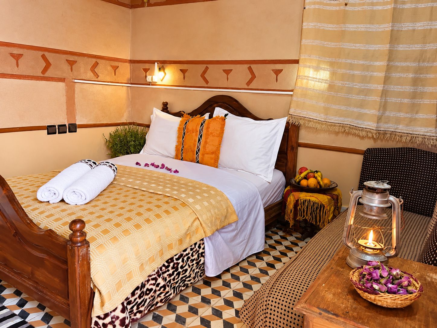 A hotel room with a checkered floor, a bed with a patterned bedspread, a nightstand with a lantern, and a chair.