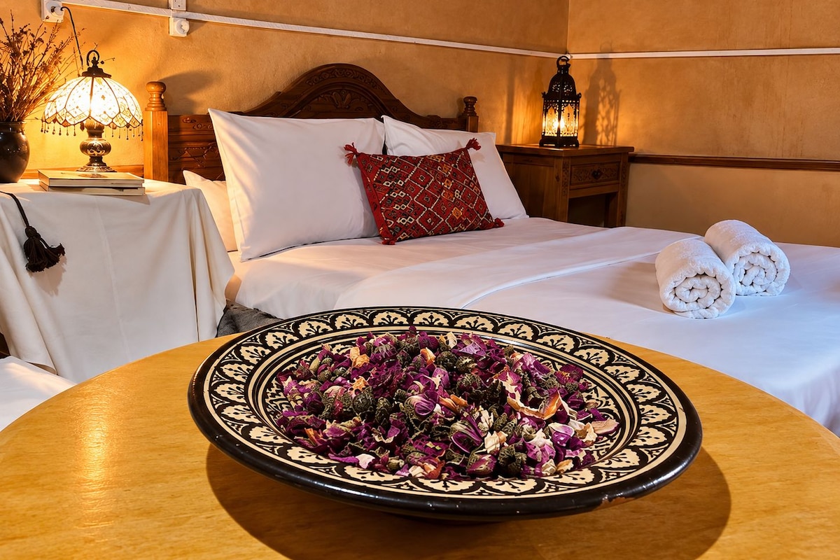 A hotel room with a bed, pillows, a bedside table with a lamp, and a decorative plate with dried flowers.
