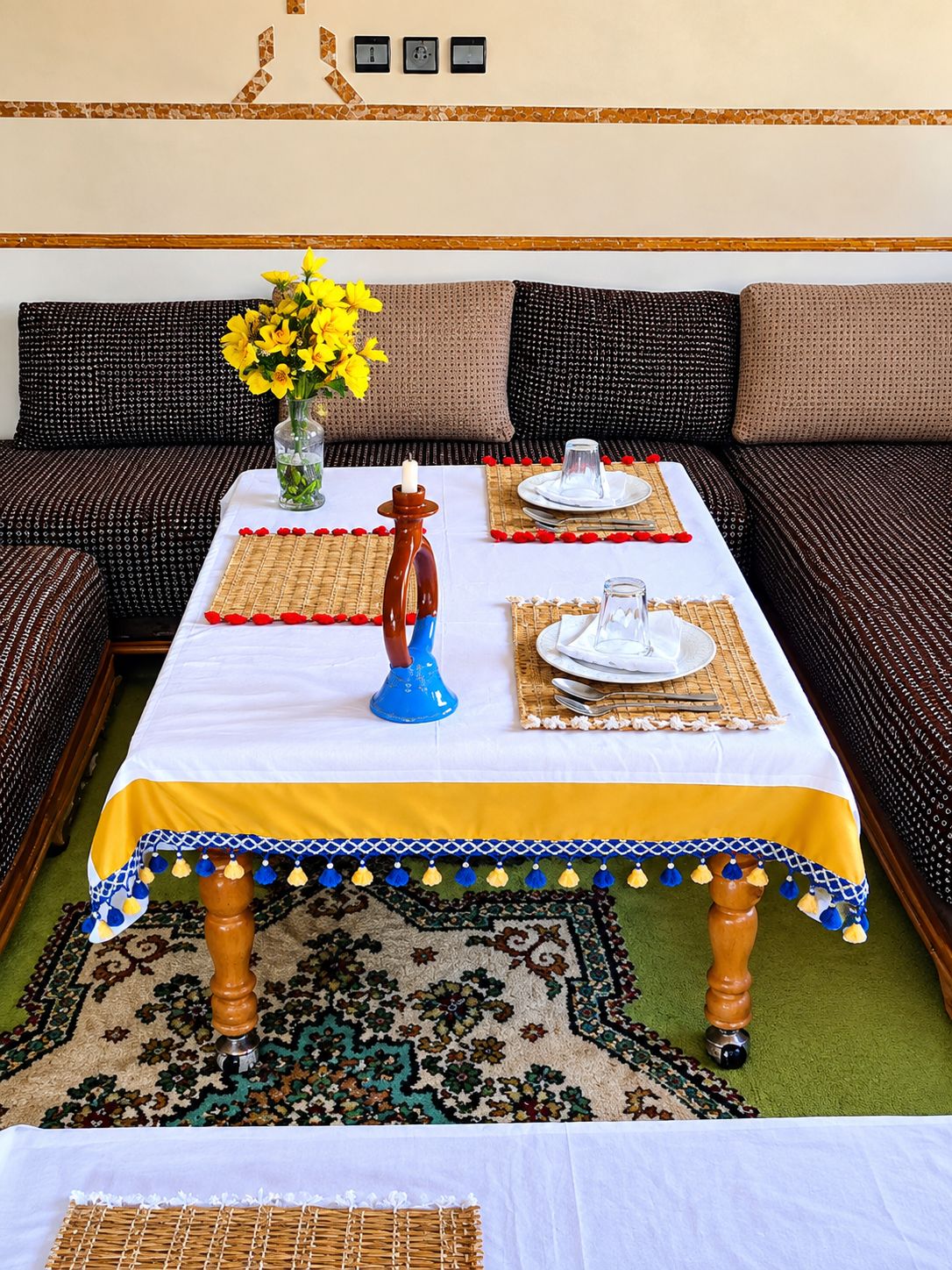 Un coin repas avec une table dressée pour deux personnes, un vase rempli de fleurs jaunes et, au fond, un canapé à motifs.