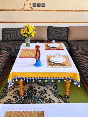 A dining area with a table set for two, a vase with yellow flowers, and a patterned sofa in the background.