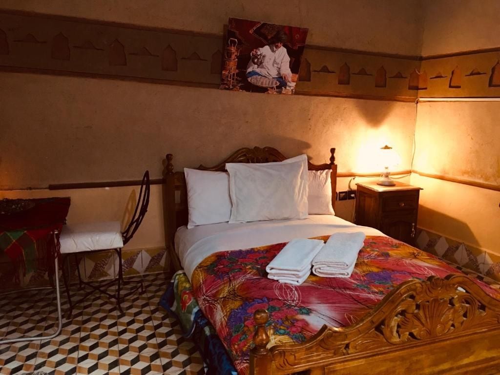 A hotel room with a checkered floor, a bed with a floral bedspread, a wooden headboard, a nightstand with a lamp, and a chair.
