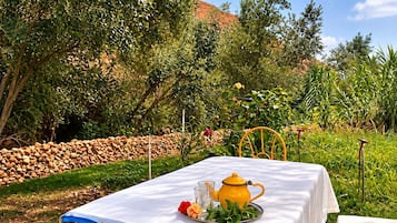 Mesa de comedor al aire libre con una tetera amarilla, vasos y una bandeja de comida, rodeada de follaje y un muro de piedra.