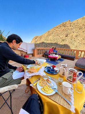 Un área de comedor al aire libre con una mesa lista para el desayuno, incluyendo platos de comida, tazas y una tetera.