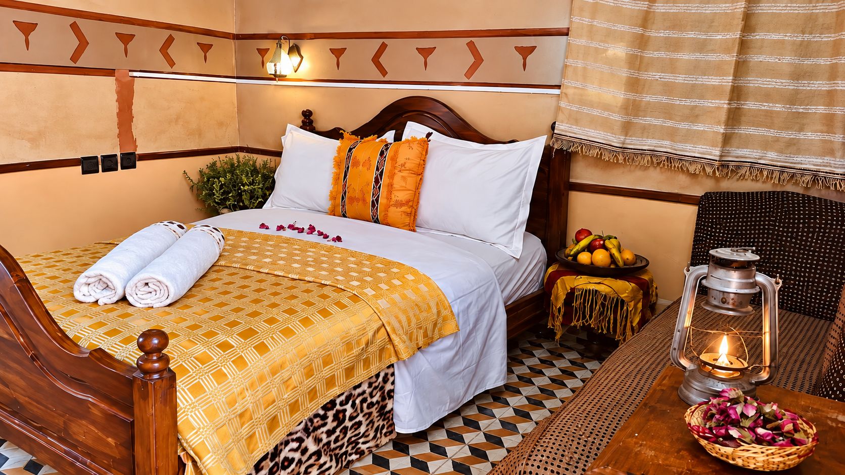 Cama com colcha estampada, travesseiros e toalhas dobradas. Pequena mesa com lanterna e frutas.