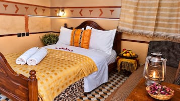 Cama com colcha estampada, travesseiros e toalhas dobradas. Pequena mesa com lanterna e frutas.