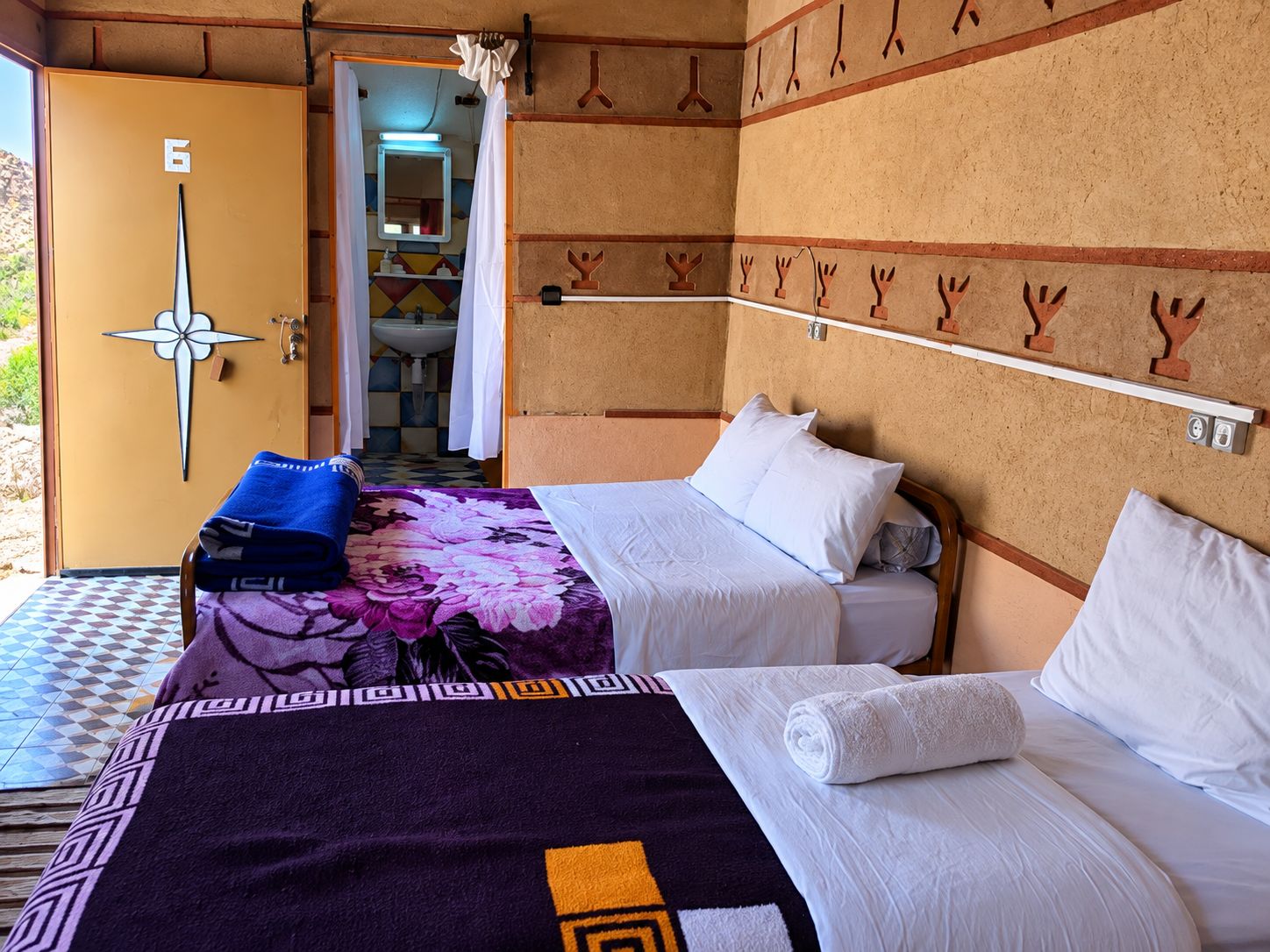 A room with two beds, a bathroom visible through an open door, and a towel rack on the wall.
