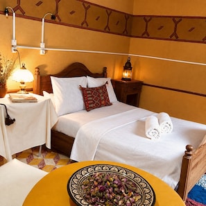 A neatly made bed with white linens and a decorative pillow, a bedside table with a lamp, and a bowl of dried flowers on a yellow table.