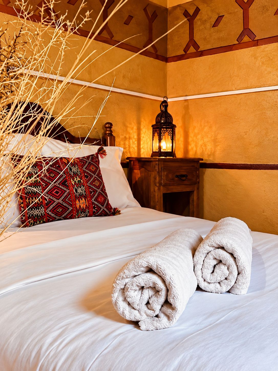 A neatly made bed with rolled towels, a patterned pillow, and a decorative wall with geometric designs.