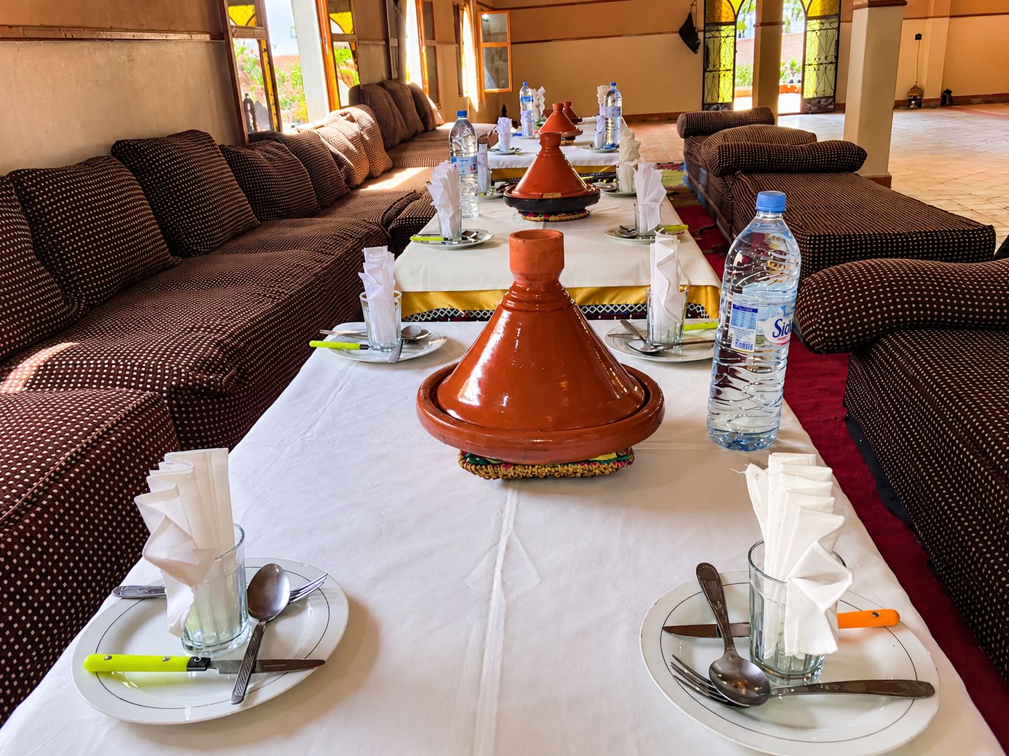 Un espace repas avec une longue table dressée pour un repas, agrémenté d’un tajine au centre, de bouteilles d’eau et de coussins traditionnels marocains.