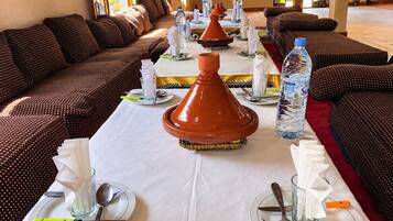 Un espace repas avec une longue table dressée pour un repas, agrémenté d’un tajine au centre, de bouteilles d’eau et de coussins traditionnels marocains.