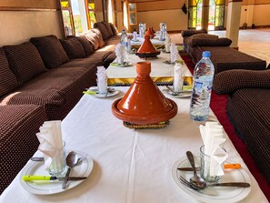 A dining area with a long table set for a meal, featuring a central tagine, water bottles, and traditional Moroccan cushions.