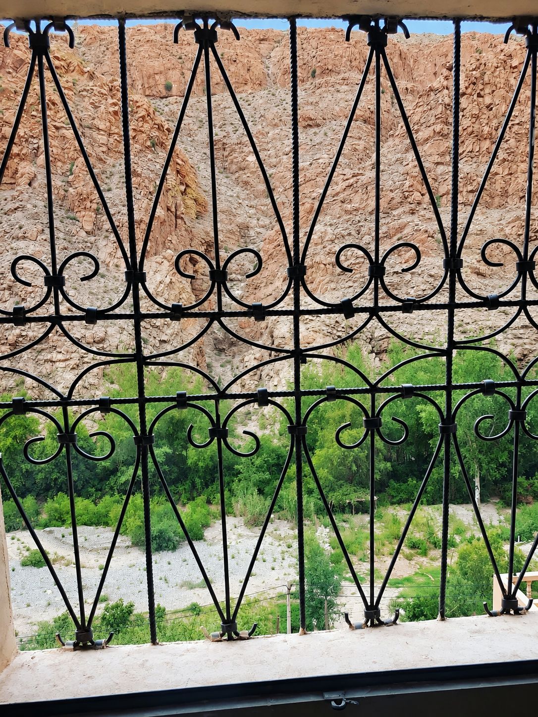 A metal gate with intricate designs, leading to a rocky outdoor area with sparse vegetation.