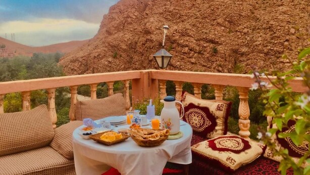 An outdoor dining area with a table set for two, surrounded by cushioned seating and a view of a mountainous landscape. - Hôtel panoramique de la vallée (Ait Sedrate Jbel El Soufla)