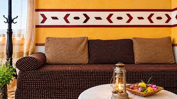 A room with a sofa set against a yellow wall, a patterned throw pillow, a table with a lantern and a fruit basket, and a window with curtains.