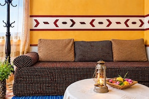 Una habitación con un sofá frente a una pared amarilla, un cojín estampado, una mesa con un farol y una cesta de frutas, además de una ventana con cortinas.