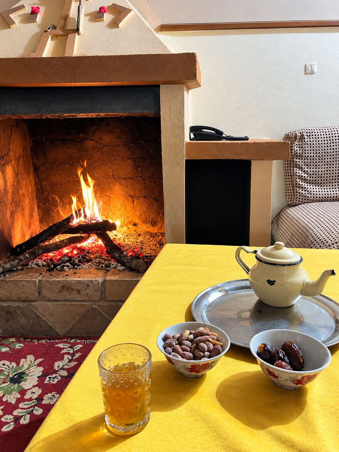 Une pièce avec une cheminée, une nappe jaune, une théière et des bols remplis de noix et de dattes.