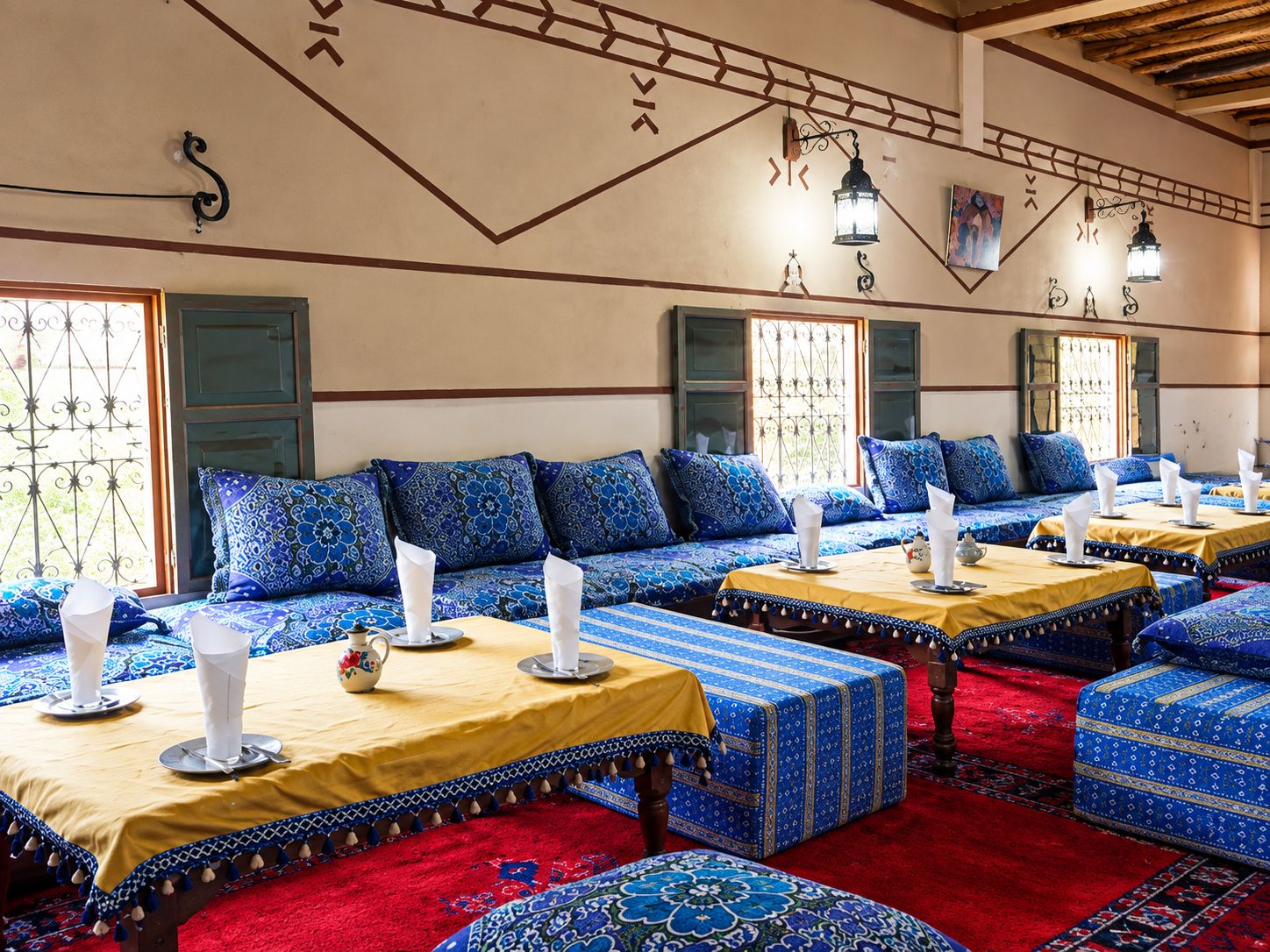 A room with multiple seating areas, each with a table set for dining, featuring patterned cushions and a red carpet.