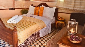 A bedroom with a checkered floor, a bed with a patterned bedspread, a bedside lamp, and a small table with a basket. - Hôtel panoramique de la vallée (Ait Sedrate Jbel El Soufla)