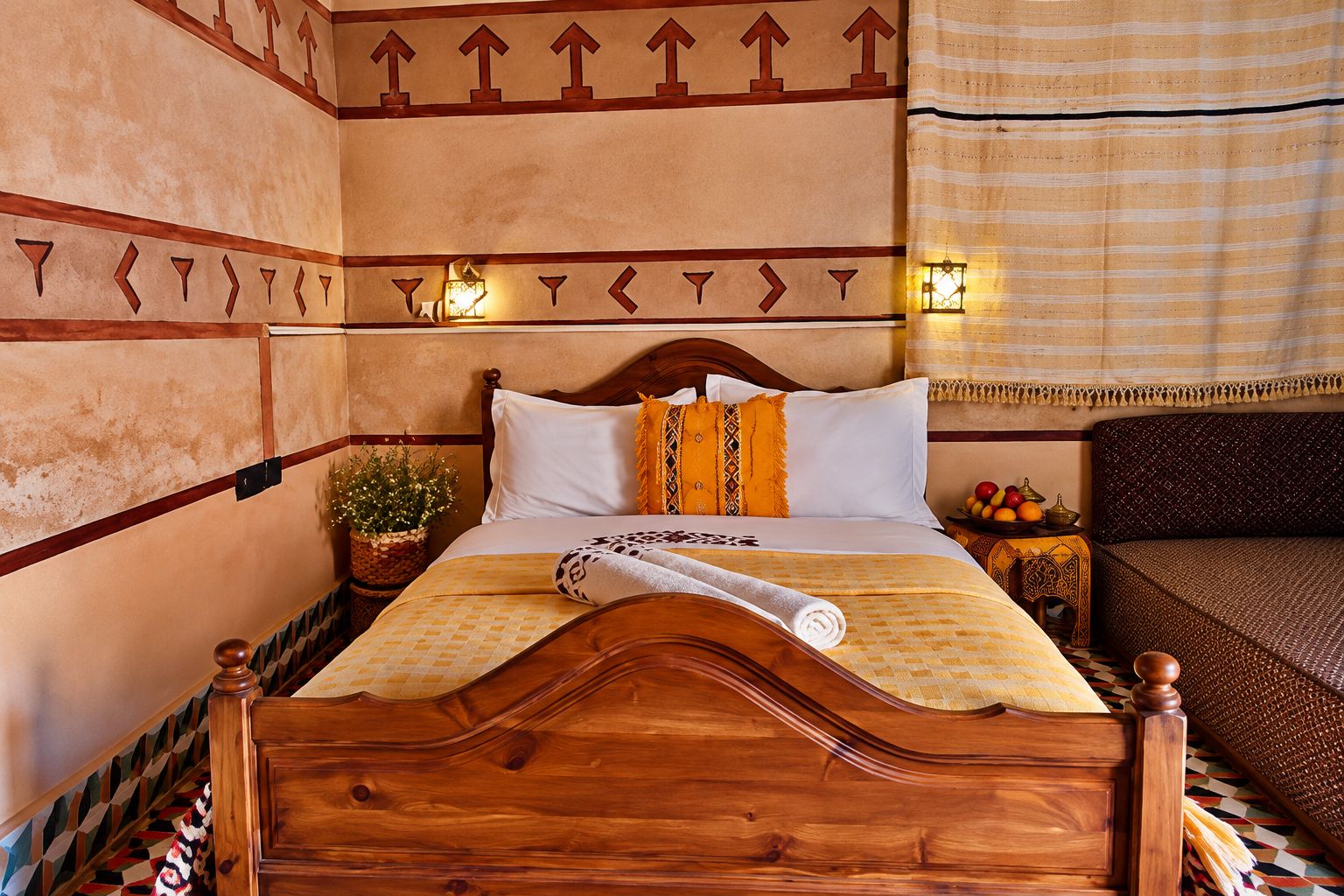 Una camera d'albergo con un letto di legno, una pianta in vaso, un comodino con una lampada e vista sull'esterno.
