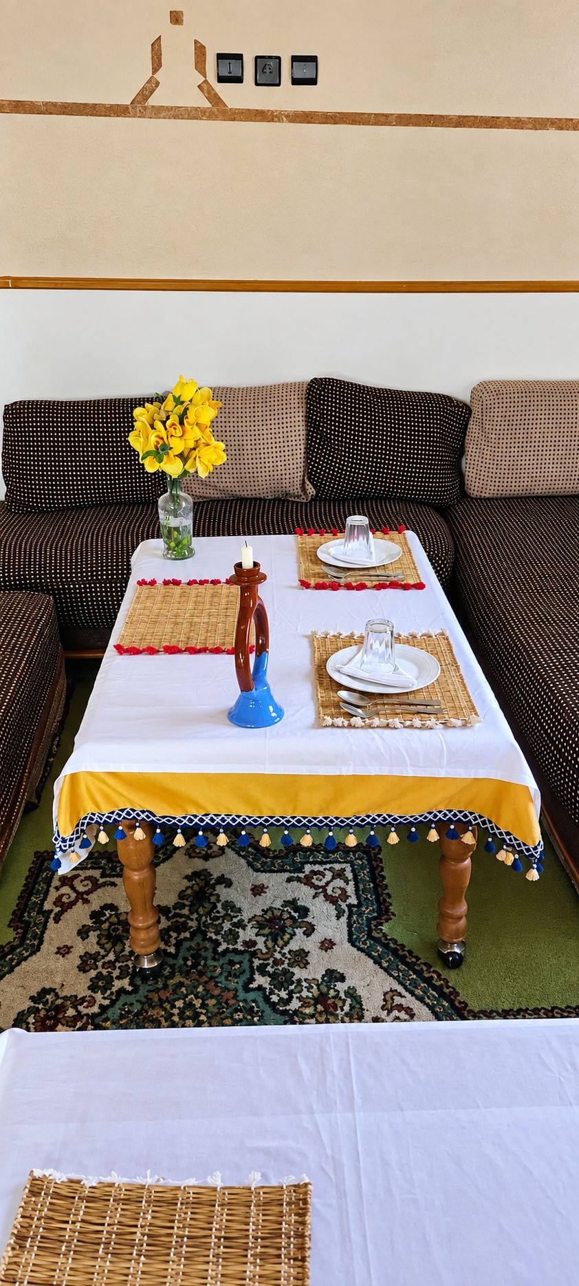 Un área de comedor con una mesa dispuesta para dos personas, un jarrón con flores amarillas y una alfombra estampada.