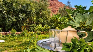 Ambiente de jardim com bule e copos em um tabuleiro, cercado por vegetação e uma formação rochosa vermelha ao fundo.