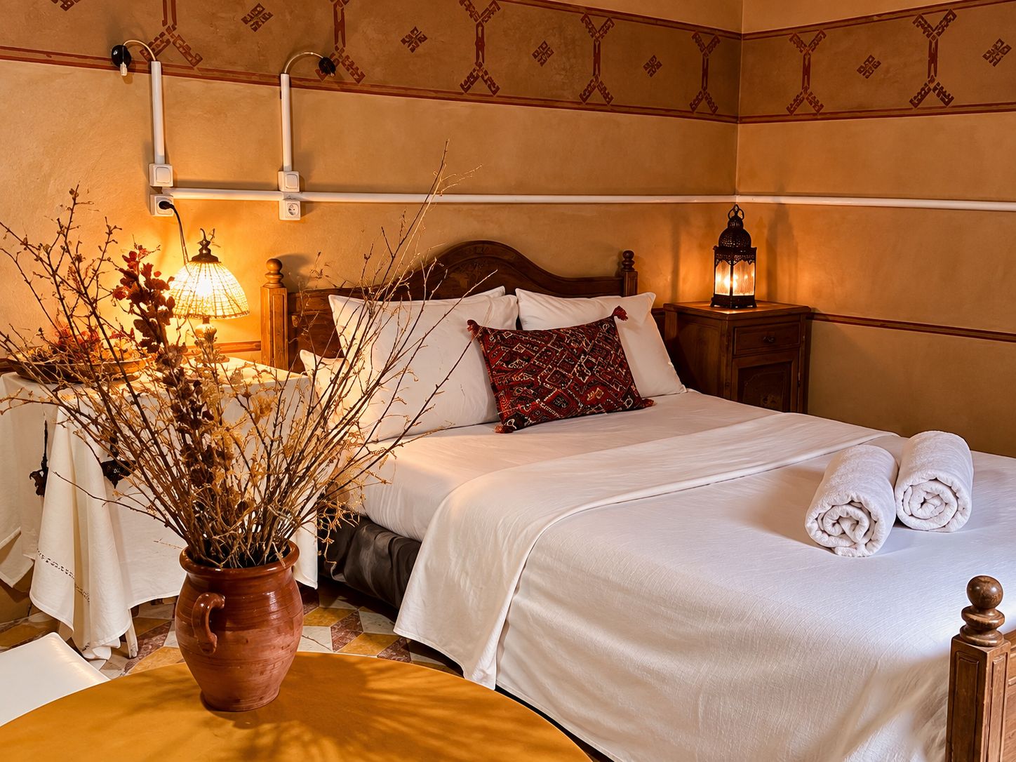 A hotel room with a bed, bedside lamps, a vase with branches, and a neatly made bed with towels.