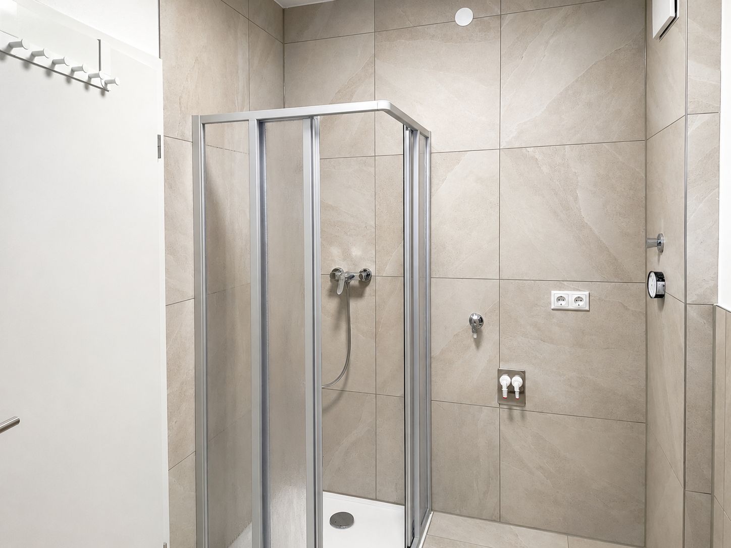 A modern bathroom with a glass-enclosed shower, beige tiles, and a visible electrical outlet.
