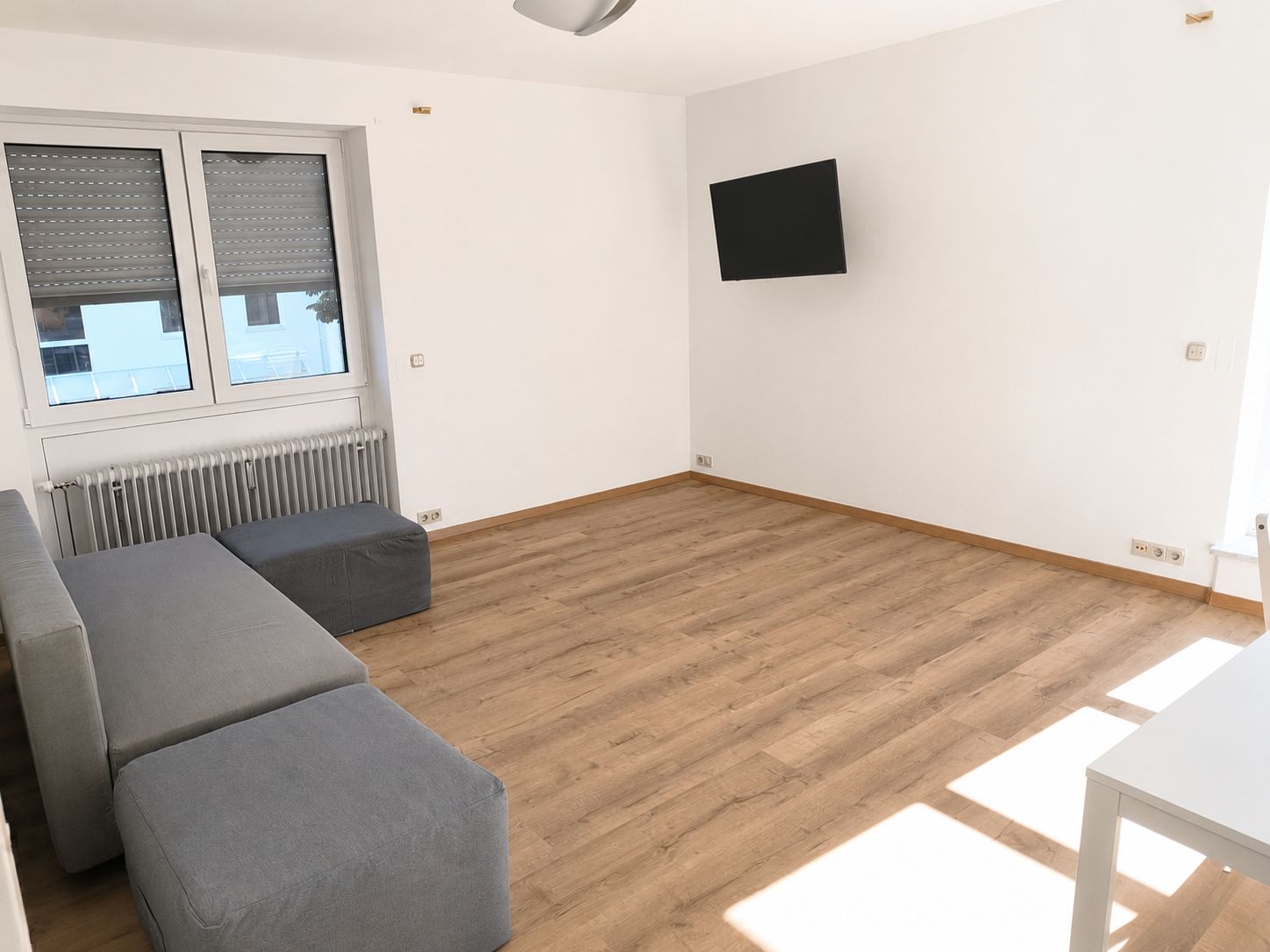 A room with a wooden floor, a grey sofa, a white table, and a TV mounted on the wall.
