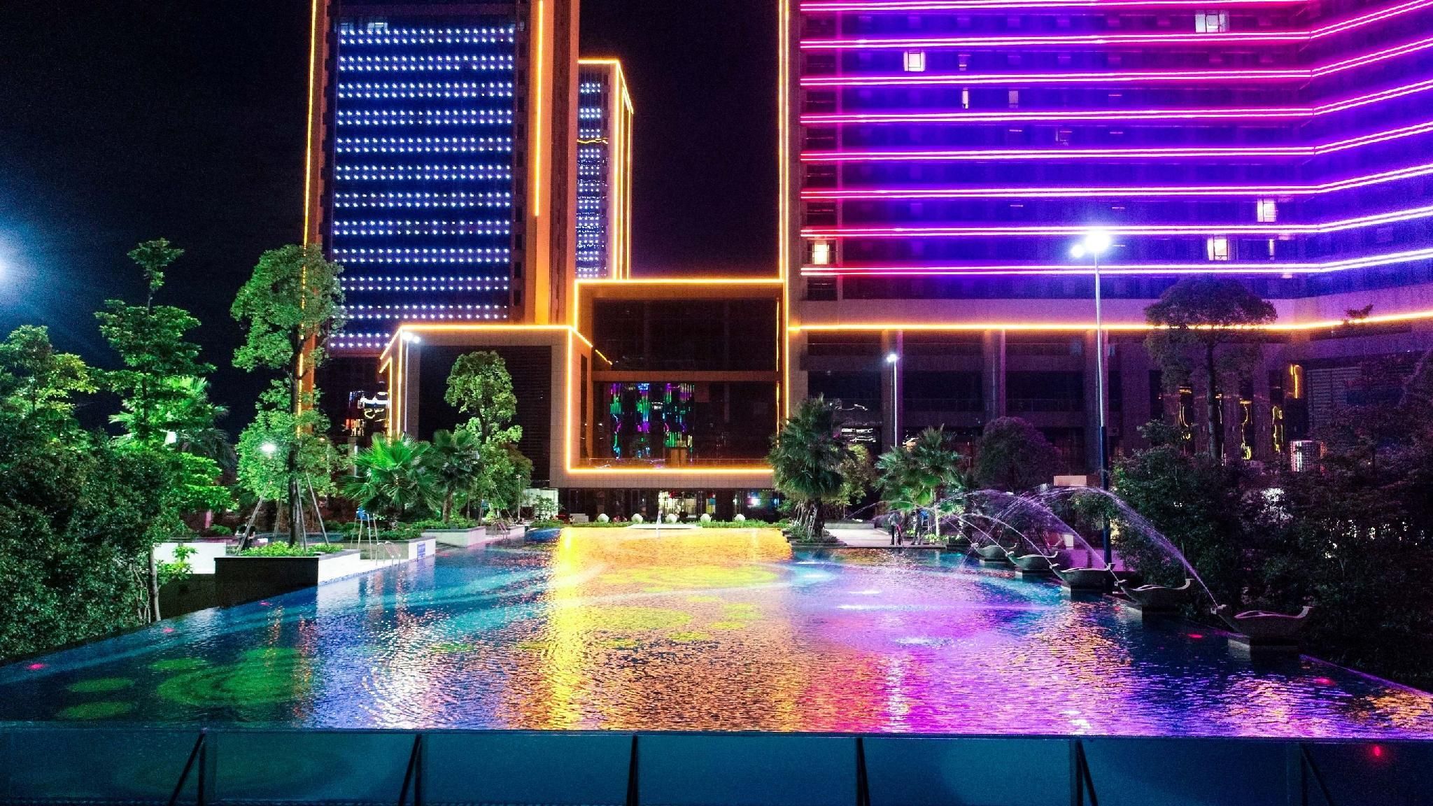 Une vue nocturne d’un hôtel avec une piscine, entouré d’arbres et illuminé par des néons.