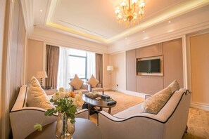 A hotel room with a sofa set, a coffee table, a flat-screen TV mounted on the wall, and a chandelier.