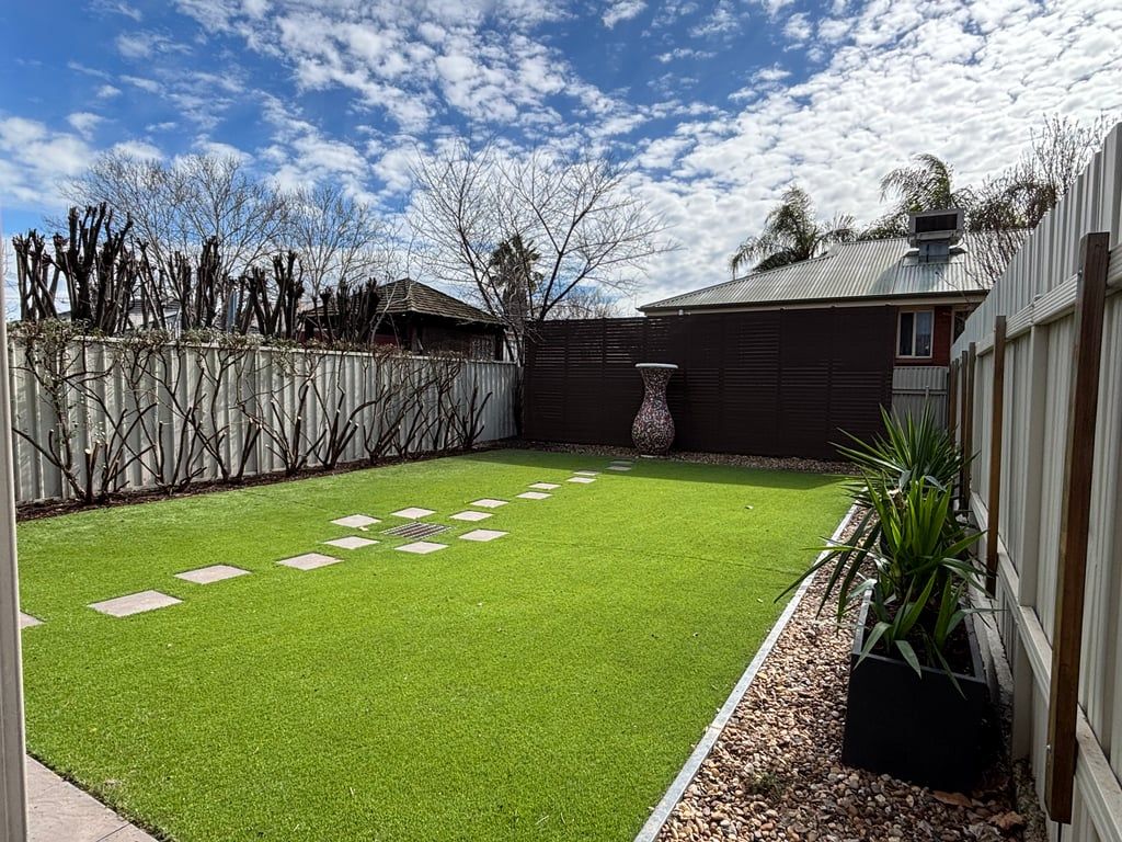 A backyard with artificial grass, a stone pathway, a wooden fence, and a house with a metal roof.
