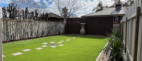 A backyard with artificial grass, a stone pathway, a wooden fence, and a house with a metal roof.