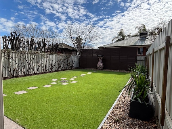 A backyard with artificial grass, a stone pathway, a wooden fence, and a house with a metal roof.