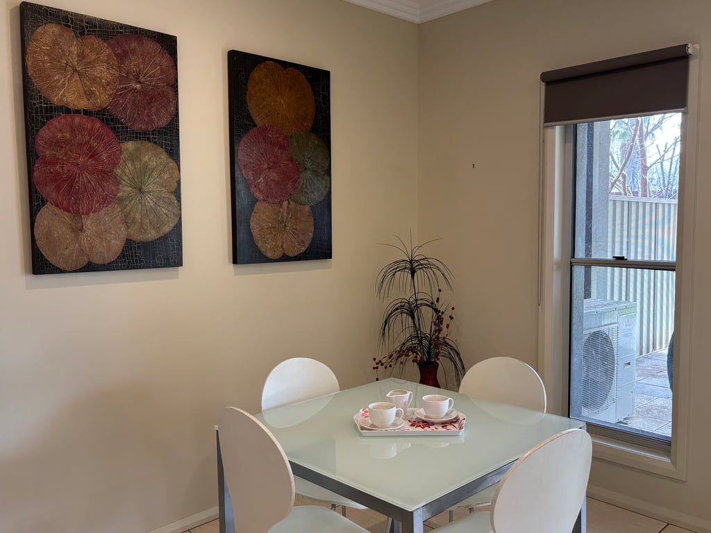 A dining area with a glass table, white chairs, and two paintings on the wall.