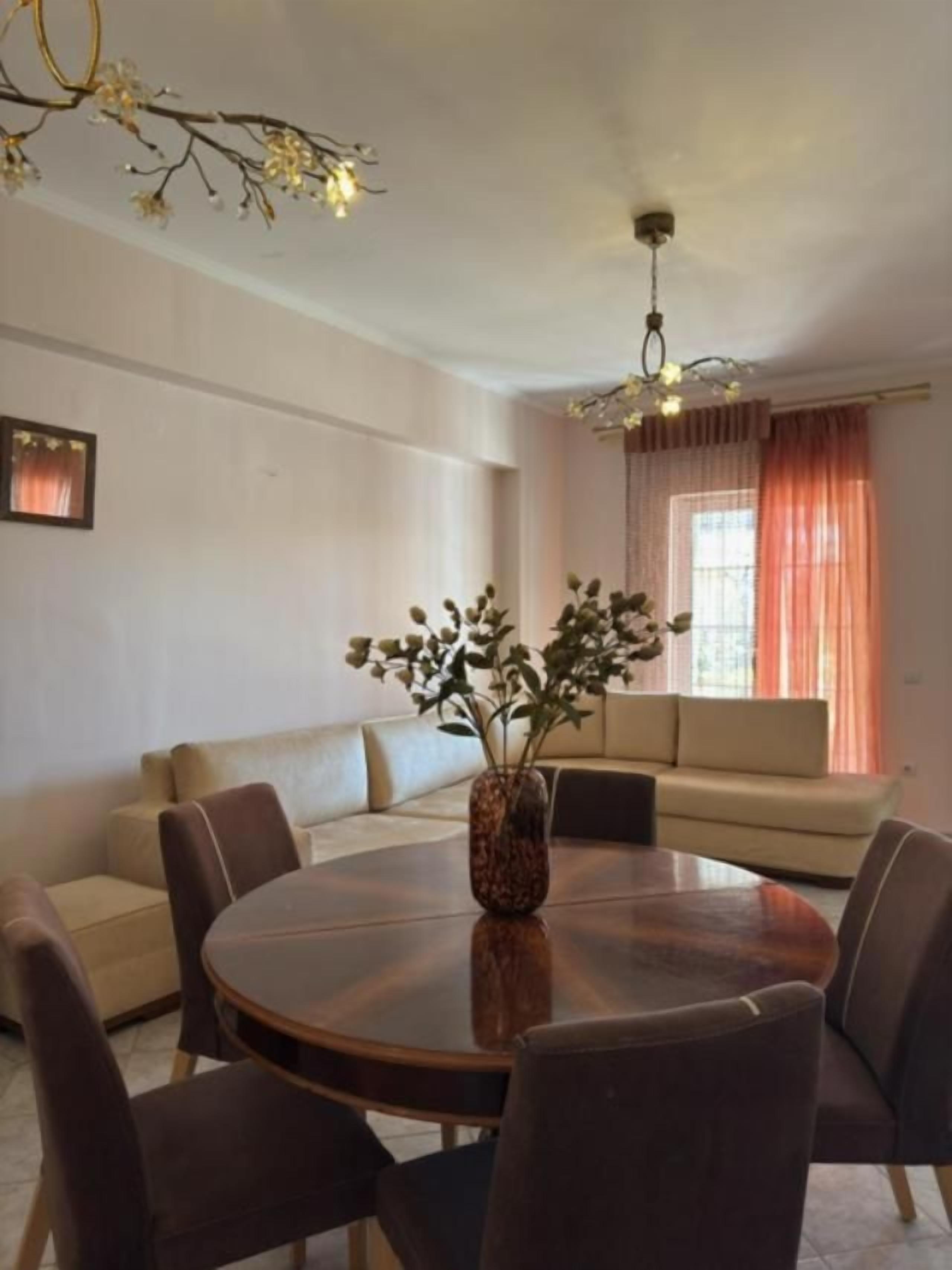 A dining area with a round table, chairs, a vase with flowers, a sofa, and a window with curtains.