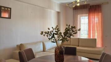 A dining area with a round table, chairs, a vase with flowers, a sofa, and a window with curtains.