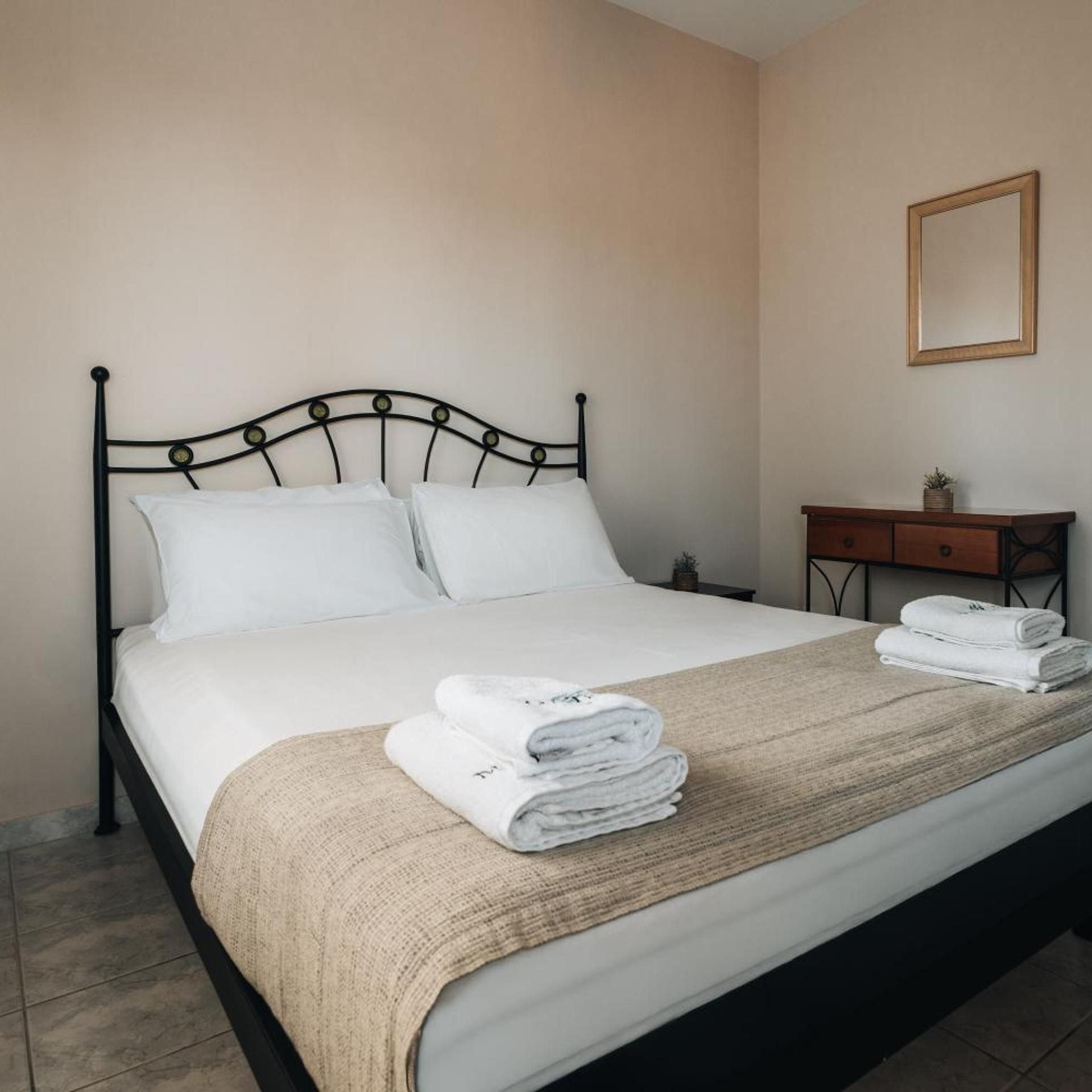 A neatly made bed with white linens and a beige throw blanket, a wooden nightstand with a mirror and a small plant, and a tiled floor.