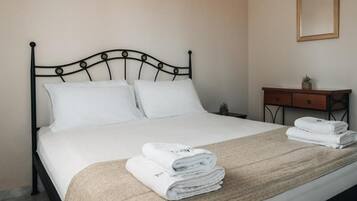 A neatly made bed with white linens and a beige throw blanket, a wooden nightstand with a mirror and a small plant, and a tiled floor.