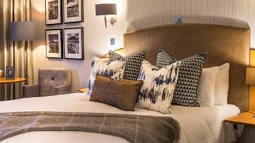 A hotel room with a large bed, multiple pillows, a bedside table with a lamp, a chair, and framed pictures on the wall.