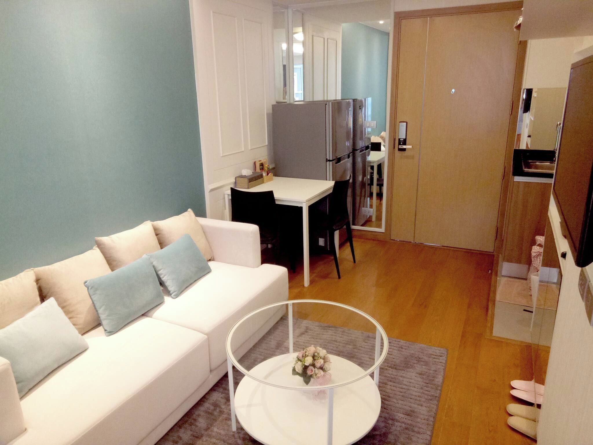 A modern living room with a white sofa, a coffee table, and a dining area with a refrigerator.