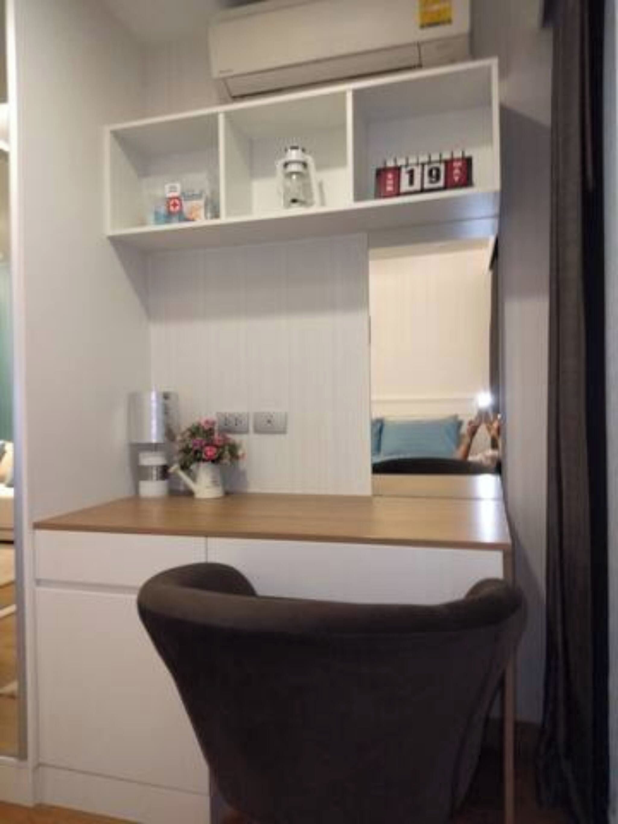 A room with a wooden desk, a chair, a shelf with items, and a bed with a person in the background.