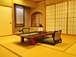 Una habitación con tatami, una mesa baja, sillas, un celosía japonés y una obra de arte enmarcada en la pared.